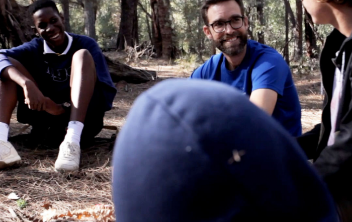 Male teacher and students sitting in a circle under some trees, smiling at each other